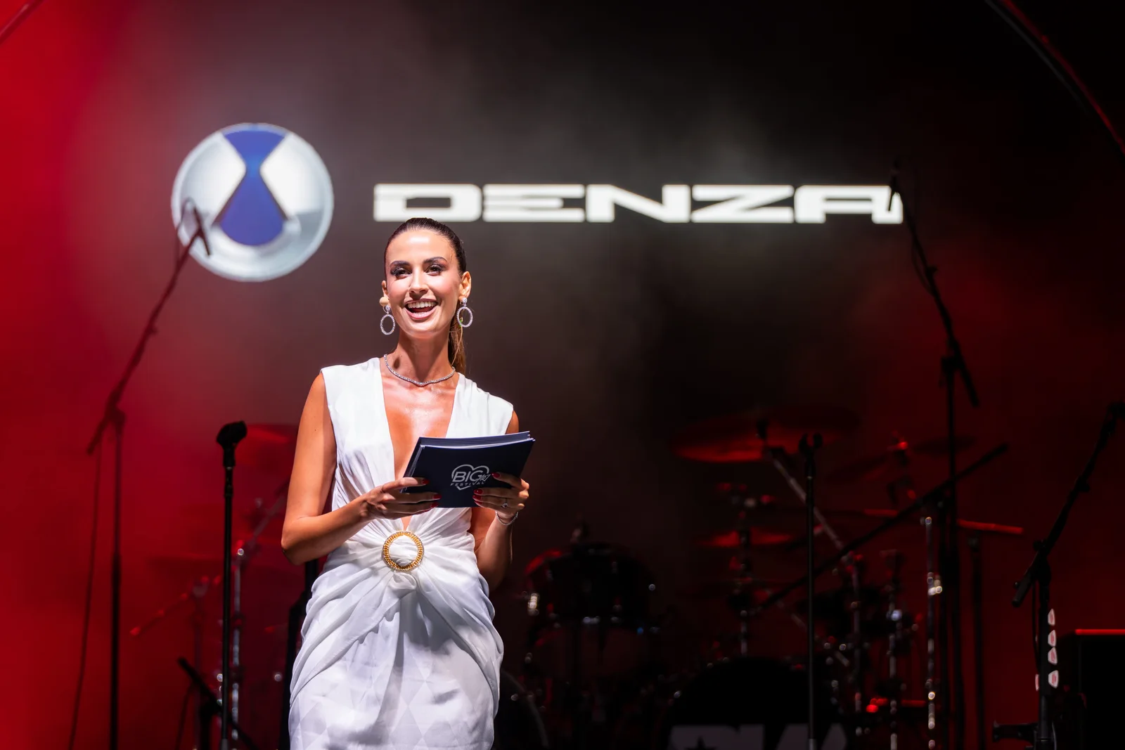 Host Eleonora Incardona on stage in a white gown beneath the DENZA logo