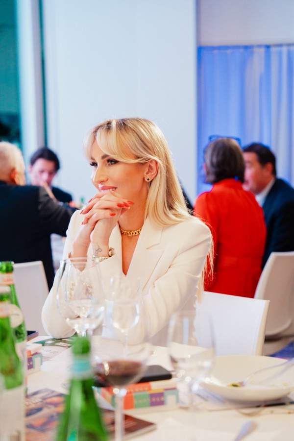 Irina Biss smiles while seated at a dinner table during a gala event
