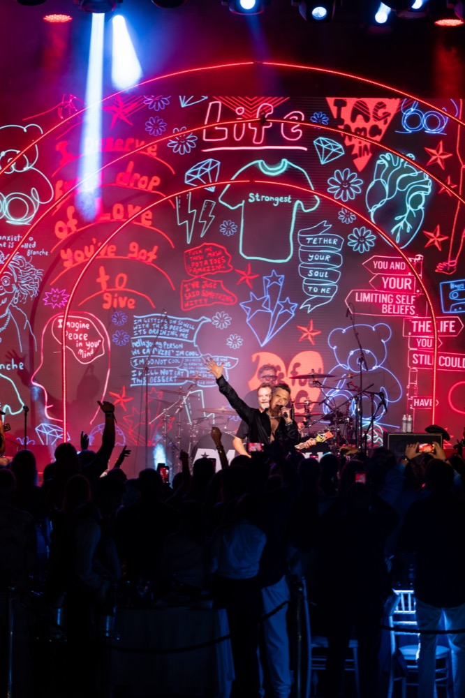 Robbie Williams performs on stage with one arm raised in front of a colorful illustrated backdrop