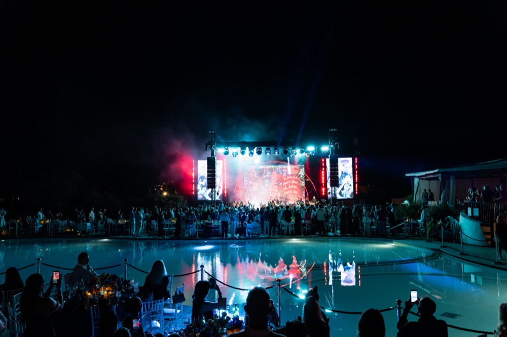 A poolside stage lit in red and blue hosts the performance as guests watch from tables around the water at night